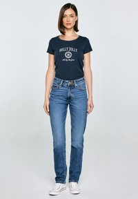 Jūras spēku T-krekls ar "HOLLY JOLLY" grafiku, saskaņots ar gaiši ziliem džinsiem. Modelis valkā baltas kedas. Vienkāršs dizains, ikdiena fit.