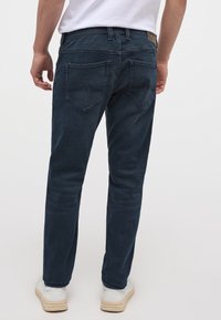 Sötétkék farmernadrág, slim fit fazonnal, két hátsó zsebbel, kontrasztos varrással és barna bőrpáttal a derékrészen.