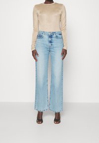 Helle blaue weit geschnittene Jeans mit hoher Taille, kombiniert mit einem beigen Langarmoberteil aus glattem Stoff. Schwarze Pumps mit offenem Zeh.