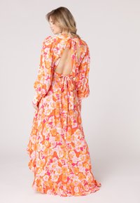 Maxi-jurk in oranje met bloemenpatronen in roze en wit. Kenmerken zijn een diepe uitsnijding op de rug, pofmouwen en een strik op de taille aan de achterkant.