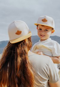 Due persone indossano berretti beige con visiera gialla e un logo marrone. Lo sfondo presenta un paesaggio montano.