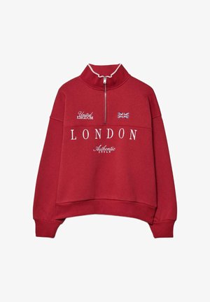 Czerwona bluza polarowa z wysokim kołnierzem, zamkiem błyskawicznym i białym haftowanym napisem: „LONDON”, „United Kingdom” oraz małym akcentem w postaci flagi Wielkiej Brytanii.