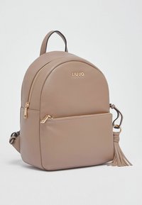 Sac à dos beige arrondi en matériau synthétique texturé. Il comporte une fermeture éclair dorée, une poche avant et un accessoire décoratif en forme de pompon.