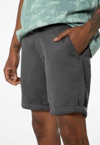 Shorts in denim grigio con orli arrotolati, tasche laterali e una texture liscia, abbinati a una T-shirt verde chiaro con stampa.