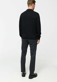 Mees seisab seljaga, seljas must pikkade varrukatega särk, tumehallad püksid ja mustad kingad heledal ühtlases taustal.