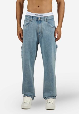 Mand iført lyseblå løstsiddende jeans, hvide sneakers og hvidt undertøj med kant med teksten "PEGADOR", stående foran en ensfarvet baggrund.