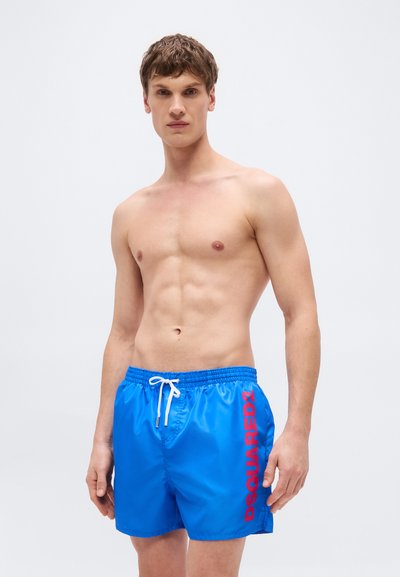 Jeune homme torse nu portant un short de bain bleu vif avec le texte rouge "DSQUARED2" sur le côté, sur un fond blanc uni.