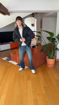 Črna puhovka z zadrgo in kapuco, obrobljeno s krznom, oblečena čez belo majico, kombinirana z modrimi jeans hlačami in belimi supergami. Notranja scena.