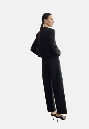 Mujer con el cabello largo y oscuro recogido en una cola de caballo, que lleva una chaqueta negra entallada, pantalones de pierna ancha y sandalias negras de tacón alto, de pie y mirando hacia la derecha.