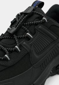 Čierna atletická obuv s textúrovaným zvrškom s ryhami, elastickými šnúrkami, výrazným logom Nike a robustným dizajnom podrážky.