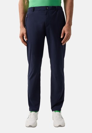 Man draagt marineblauwe casual broek met zijritszakken, groene shirt en witte sneakers tegen een effen achtergrond.