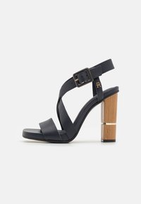 Tummansininen nahkainen korkokenkä sandaali, jossa on leveät ristiin menevät hihnat, säädettävä solki ja paksu puinen lohkare korko, jossa on kultaiset yksityiskohdat.