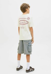 T-shirt crème clair avec un texte graphique rouge et un logo au dos, associé à un short cargo bleu avec des poches latérales. Chaussures marron et chaussettes blanches.