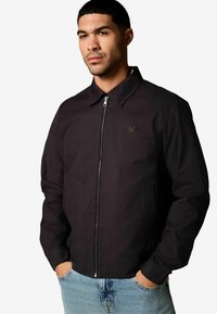 Schwarze Jacke mit Reißverschluss, Kragen und seitlichen Taschen, die ein dezentes goldenes Logo auf der linken Brust trägt. Der Stoff hat eine glatte Textur.