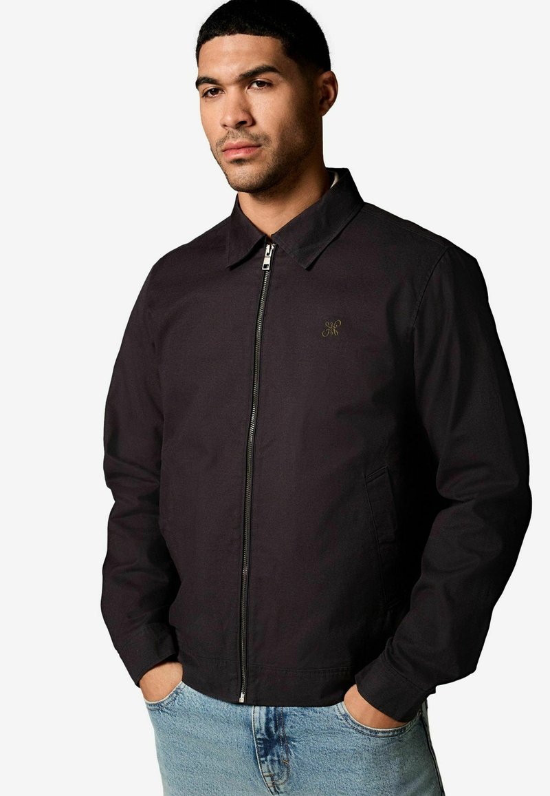 Schwarze Jacke mit Reißverschluss, Kragen und seitlichen Taschen, die ein dezentes goldenes Logo auf der linken Brust trägt. Der Stoff hat eine glatte Textur.