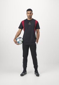 Juodas sportinis marškinėliai su raudonais akcentais ir auksiniu logotipu, derinami su juodomis treniruočių kelnėmis ir juodais futbolo batais, laikant daugiaspalvį futbolo kamuolį.