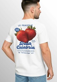 Weißes T-Shirt mit einem Grafik von zwei roten Erdbeeren auf dem Rücken, mit blauer Schrift, die "Della Calabria" und "LE FRAGOLE" liest.