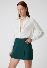 Weißes Langarmshirt mit V-Ausschnitt und Knopfdetails, kombiniert mit einem türkisfarbenen Minirock mit seitlicher Faltendetails. Glatte Stoffoberfläche und taillierte Passform.