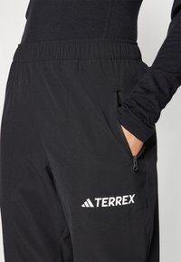 Crne Terrex hlače imaju elastičnu navlaku, bočni džep sa zatvaračem i bijeli logo na bedrima. Mekani, glatki materijal s minimalnom teksturom.