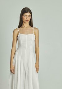 Weißes Leinenkleid mit tailliertem Oberteil, quadratischem Ausschnitt und dünnen Trägern. Der fließende Rock hat vertikale Nähte und ein plissiertes Design.