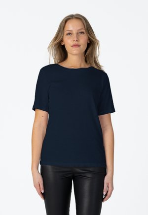 Femme aux longs cheveux blonds portant une chemise à manches courtes bleu marine et un pantalon en cuir noir, debout sur un fond blanc.