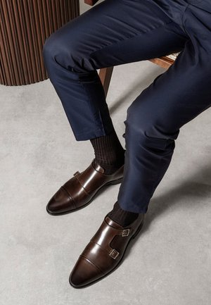 Bruine leren dubbele monk strap schoenen met een gepolijste afwerking, gedragen met zwarte sokken en navy broek op een lichtgrijze vloer.
