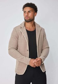 Homme avec des cheveux courts et bouclés et une barbe, portant un blazer texturé beige par-dessus une chemise noire, se tenant devant un fond clair uni.