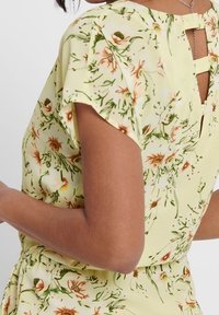Robe jaune à motif floral avec manches courtes, une ouverture en forme de goutte dans le dos, et un tissu texturé avec des accents verts et orange.