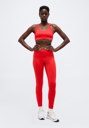 Persona atletica in piedi con le mani sui fianchi, indossa un reggiseno sportivo rosso, leggings rossi, calzini bianchi e scarpe da ginnastica bianche su uno sfondo uniforme.