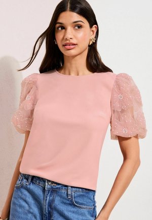 Jeune femme portant un chemisier rose avec des manches bouffantes transparentes brodées de fleurs et un jean en denim bleu, debout devant un fond uni.