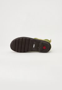 Suela de zapatilla verde con una suela exterior de goma negra texturizada, que presenta patrones de agarre en zigzag y un acento rojo cerca del talón.