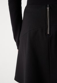 Jupe noire en tissu extensible, dotée d'une fermeture éclair en métal argenté visible, texture lisse et design ajusté avec des détails cousus.