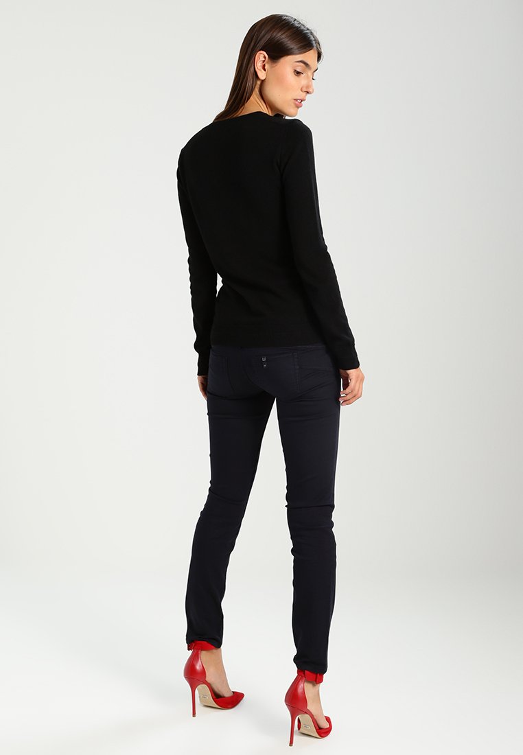 Schwarzer Pullover mit langen Ärmeln im taillierten Stil, kombiniert mit dunkelblauen Slim-Fit-Hosen und roten High Heels. Schlichtes Design, glatte Textur.