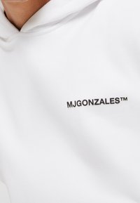 Sweat à capuche blanc en tissu doux, avec un petit logo noir "MJGONZALES™" imprimé sur la poitrine. Design simple et épuré avec une capuche à cordon.