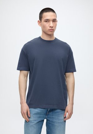 Bărbat cu păr scurt și închis la culoare, purtând un tricou simplu albastru închis și blugi deschiși la culoare, stând în fața unui fundal alb.