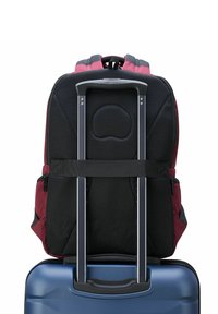 Mochila roja y negra con parte trasera de malla, montada sobre una maleta dura azul. Cuenta con un mango extensible y una correa resistente.