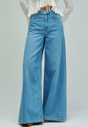 Femme portant un jean bleu taille haute à jambes larges, associé à un chemisier court blanc en dentelle à manches longues et des sandales.