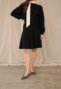 Moteris vilki juodą ilgomis rankovėmis suknelę ir baltą šaliką, stovi ant pilkos grindų dangos su smėlio spalvos užuolaidomis fone.