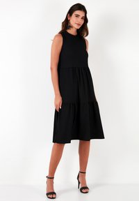 Black sleeveless dress with a fitted bodice and a tiered skirt. Smooth fabric with a textured appearance, falling knee-length.