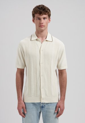 Jonge man met krullend haar, die een kort mouw crème overhemd met knopen van textiel draagt en een lichtblauwe spijkerbroek, tegen een grijze achtergrond.