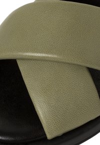 Sandales en cuir texturé vert olive avec de larges brides entrecroisées sur une semelle noire, présentant une finition lisse et mate et un design minimaliste.