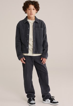 Donkergrijze corduroy jas met knopen, bijpassende corduroy broek en zwarte sneakers met grijze accenten. Witte t-shirt eronder.