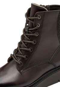 Barna bőr bokacsizma texturált díszítéssel, kerek orral és kontrasztos fűzőrendszerrel. Fém szemekkel és sima felülettel rendelkezik.