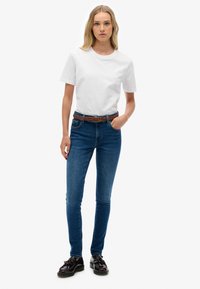 Una camiseta blanca de manga corta combinada con un jeans ajustado de color azul oscuro, acentuada por un cinturón trenzado marrón y zapatos negros con detalles de borlas.