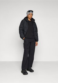 Doudoune noire avec capuche, associée à un t-shirt noir à manches longues et un pantalon cargo. Bonnet en tricot gris et baskets noires avec des accents.