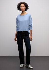 Svetle modrý svetr volného střihu a širokým výstřihem, doplněný tmavými džíny s rovnými nohavicemi a stříbrnými teniskami. Celý outfit má hladkou texturu.