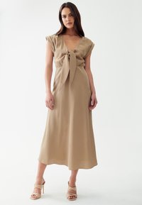 Robe midi beige avec des manches courtes, détail de ceinture à nouer, tissu lisse et silhouette fluide. Associée à des chaussures à talons beiges à lanières.