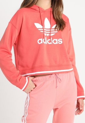 Vrouw die een koraalrode cropped hoodie van Adidas draagt met een wit logo en bijpassende roze Adidas trainingsbroek met witte strepen en een taille met trekkoord.