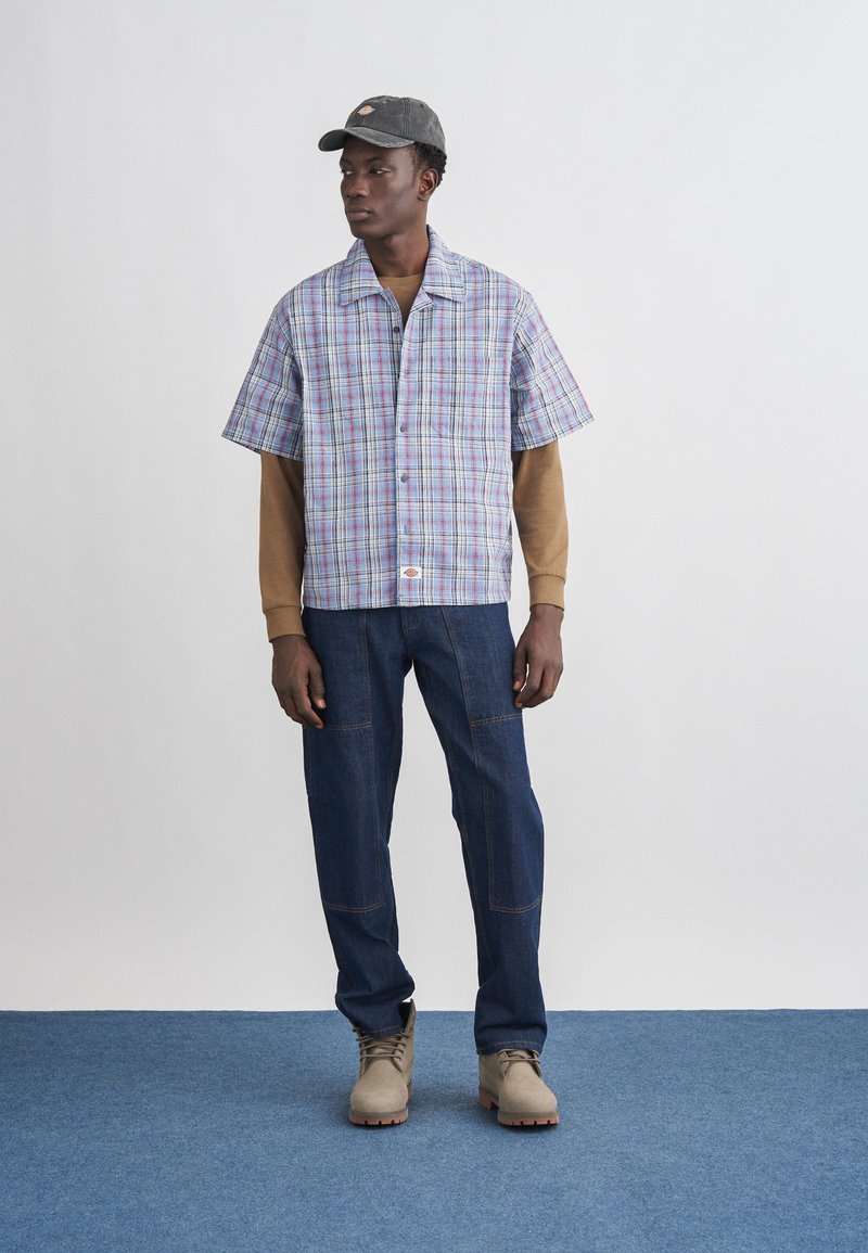 Mann mit schwarzer Kappe, hellblauem kurzärmligem karierten Hemd über braunem Langarmshirt, dunkelblauer Jeans und beigefarbenen Stiefeln, stehend auf blauem Teppich.