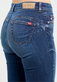 Vaqueros de mezclilla azul oscuro con un diseño ajustado, que cuentan con un bolsillo trasero cosido, etiqueta con el logo y ligeros detalles de desgaste en la tela.